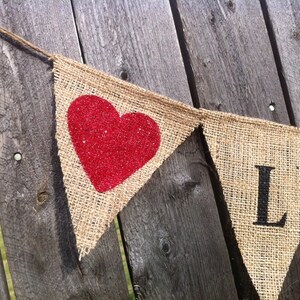 May include: A burlap banner with a red glitter heart and the letter "L" in black. The banner is strung on a twine string and is hanging on a wooden fence.