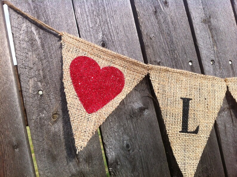 May include: A burlap banner with a red glitter heart and the letter "L" in black. The banner is strung on a twine string and is hanging on a wooden fence.