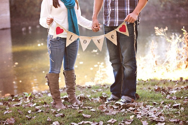 May include: A couple holding a burlap banner with red hearts and the word "LOVE" in black letters. They are standing in a grassy area with a body of water in the background.