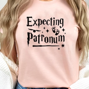 May include: A light pink t-shirt featuring black text that reads Expecting Patronum. The design includes a small winged ball, baby footprints, a lightning bolt, a wand, and star accents. The shirt is worn with a white cardigan and blue jeans.