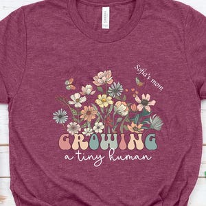 Può includere: Una t-shirt a maniche corte color bordeaux con una grafica di fiori di campo colorati e farfalle. Il testo recita Sofia's mom e GROWING a tiny human in un carattere retrò. La maglietta è presentata con scarpe da ginnastica bianche e tulipani rosa.