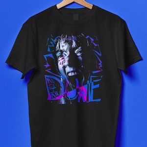 Può includere: T-shirt nera con un design grafico di un volto che urla, con accenti blu, viola e rosa. La parola "DUNE" è scritta in un font in stile graffiti sul design. La t-shirt è appesa a una gruccia di legno.