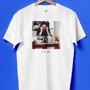 Puede incluir: Camiseta blanca con un gráfico de una mujer con las manos en la cabeza, con el texto "I'm ok" debajo. El gráfico es un collage de dos fotos de la mujer.