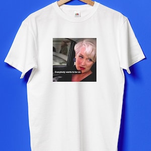 Puede incluir: Camiseta blanca con un gráfico de Meryl Streep y el texto "Everybody wants to be us". La imagen muestra un primer plano de la actriz en un coche. La camiseta está colgada en una percha de madera sobre un fondo azul.