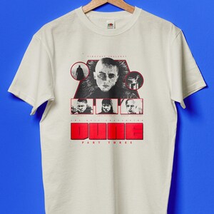Può includere: T-shirt bianca con grafica del film "Dune: Part Three". Il design include ritratti di personaggi e la parola "DUNE" in lettere rosse. La maglietta è appesa a una gruccia di legno.