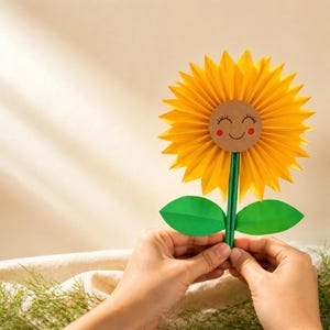 May include: A paper sunflower craft featuring a bright yellow pleated paper flower head with a circular brown center. The center has a hand-drawn smiling face with closed eyes and red cheeks. A green paper stem supports two green paper leaves.