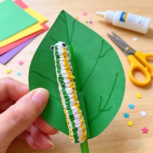 Op de afbeelding: Een handgemaakte rups bestaande uit witte, gele en groene segmenten met kleine zwarte oogjes, bevestigd aan een groen papieren blad met handgetekende nerven. De rups is gemonteerd op een groen plastic rietje voor een beweegbaar effect.