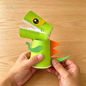 May include: A green paper dinosaur craft with a movable head and mouth. The dinosaur features white teeth, a yellow eye, and orange and green details. The craft is held by two hands on a wooden surface.