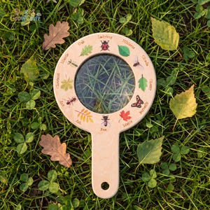 Peut inclure: Une loupe ronde en bois avec un manche présentant des illustrations imprimées et des étiquettes d'insectes et de feuilles, notamment une coccinelle, une mante, une fourmi, une abeille, un papillon, une cigale et divers types de feuillages.