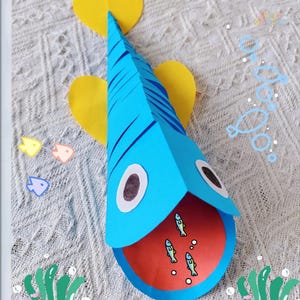Puede incluir: Un pez de papel azul hecho a mano con rayas negras y ojos grandes. El pez tiene una boca roja con tres peces pequeños en su interior. Dos aletas amarillas en forma de corazón están unidas al pez. Burbujas y peces decorativos en el fondo.