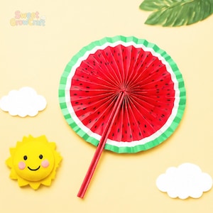 Puede incluir: Un abanico de papel circular diseñado como una rodaja de sandía con colores rojo, negro y verde. El abanico tiene un mango rojo y se acompaña de un recorte de sol amarillo con cara sonriente y dos recortes de nubes blancas.