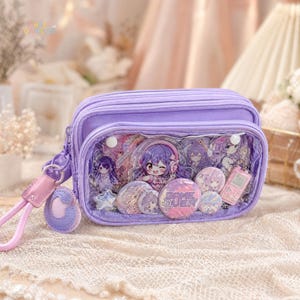 Op de afbeelding: Een lavendelkleurige etui met een transparant voorpaneel met anime-achtige karakterspelden, een kleine roze telefoon en een "GAME OVER"-knop. De etui heeft een bijpassende polsband met een decoratieve bedel.