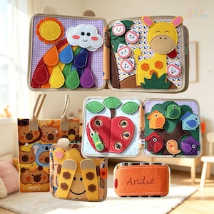 Puede incluir: Una colección de libros de fieltro coloridos y una bolsa a juego. Los libros presentan elementos interactivos como un arcoíris con gotas de lluvia, una jirafa, una manzana con una oruga y pájaros. Un estuche de fieltro naranja lleva el nombre Andie.