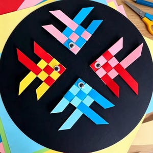 May include: Four woven paper fish shapes arranged in a circle on a black circular board. Each fish is constructed from interlocking strips of colored paper in pink, blue, red, and yellow, and features a single small plastic googly eye.