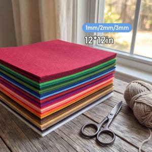 Op de afbeelding: Een stapel vierkante viltvellen in diverse kleuren waaronder rood, groen, blauw, oranje, geel, bruin en wit. De tekst 1mm/2mm/3mm en 12*12in is zichtbaar. Een zwarte metalen schaar en een bol beige garen liggen ernaast.