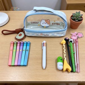 May include: A collection of stationery items including pens, a pencil case, and decorative pens. The light blue pencil case has a clear front with a cat graphic and the words "LUCKY KITTEN". Several colorful pens and a multi-color pen are also visible.