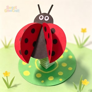 May include: A 3D paper craft ladybug with a black head, two antennae, and two red wings featuring black circular spots. The ladybug is mounted on a green circular base decorated with yellow dots, small paper flowers, and green grass blades.