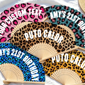 Può includere: Cinque ventagli pieghevoli con stecche in legno e foglie in tessuto con motivi leopardati in rosa, verde acqua, marrone, blu e giallo. Ogni ventaglio mostra testo bianco, tra cui "YOUR CUSTOM TEXT," "AMY'S 21ST BIRTHDAY," e "PUTO CALOR."