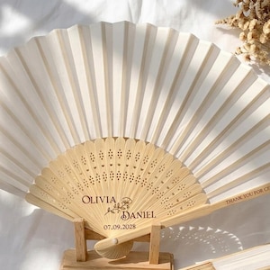 Può includere: Un ventaglio di carta bianca con manico e cornice in legno, appoggiato su un supporto in legno. Il ventaglio è aperto, mostrando il suo design a pieghe. Il manico è inciso con i nomi "Olivia & Daniel" e la data "07.09.2028".
