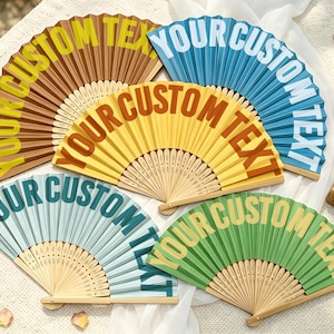May include: Five folding hand fans with wooden ribs and pleated fabric in brown, blue, yellow, light blue, and green. Each fan features the text YOUR CUSTOM TEXT printed in a contrasting color across the surface of the fabric material.