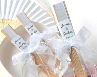 May include: White paper fans with wooden handles, tied with white lace and ribbon. Each fan has a label that reads "James & Sophia July 17, 2008". Several other white fans are visible in the background.