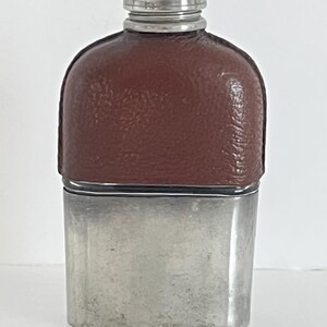 Puede incluir: Una petaca rectangular de metal con una base plateada lisa y una sección superior recubierta de cuero marrón. La petaca está rematada con un tapón de rosca plateado a juego que presenta un patrón texturizado en el borde de la tapa.