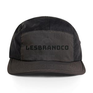 May include: Black five-panel cap with a curved brim. The front panel features the text "LESBRANDCO" in bold, black letters. The cap has a matte finish and a tonal design, with a darker shade on the brim.