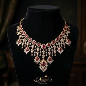 Pode incluir: Um colar dourado com pedras preciosas ovais vermelho rubi e detalhes claros e brilhantes. O colar apresenta um design em cascata com vários pingentes e é exibido em um manequim preto. A marca "Noore Jewellers" é visível.