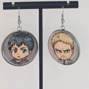 May include: A pair of round silver-toned dangle earrings featuring circular glass cabochons. Each earring displays a different anime-style character illustration: one with short dark hair and a brown jacket, and the other with short blonde hair and a suit.