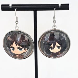 May include: A pair of round dangle earrings featuring chibi-style anime character illustrations. One earring displays a character with short brown hair, and the other shows a character with short black hair. Both are set in silver-toned metal frames.