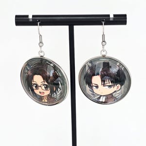 May include: A pair of round silver-toned dangle earrings featuring chibi-style anime character illustrations under a clear glass dome. One earring depicts a character with dark hair and glasses, while the other shows a character with dark hair and a scarf.