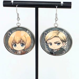 May include: A pair of round silver-toned dangle earrings featuring chibi-style anime character illustrations. The left earring depicts a character with blonde hair, and the right earring shows a character with short blonde hair and a green cape.