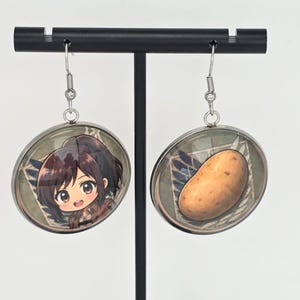 May include: A pair of round dangle earrings featuring silver-toned metal frames and hook wires. One earring displays a chibi-style anime character with brown hair, while the other earring features a single brown potato against a wing emblem background.