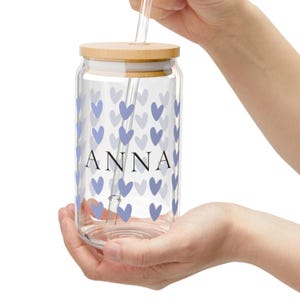 Könnte beinhalten: Ein durchsichtiger Glasbecher mit Holzdeckel und klarem Strohhalm. Der Becher ist mit einem Muster aus hellblauen Herzen und dem Namen "ANNA" in schwarzen Buchstaben verziert. Der Becher wird von einer Person gehalten.