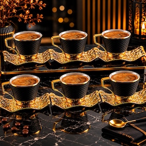 May include: Six black coffee cups with gold trim and handles, filled with coffee, sit on gold-colored saucers. The cups have a textured pattern. The saucers are placed on a black tray. A gold spoon and a black napkin are in the foreground.
