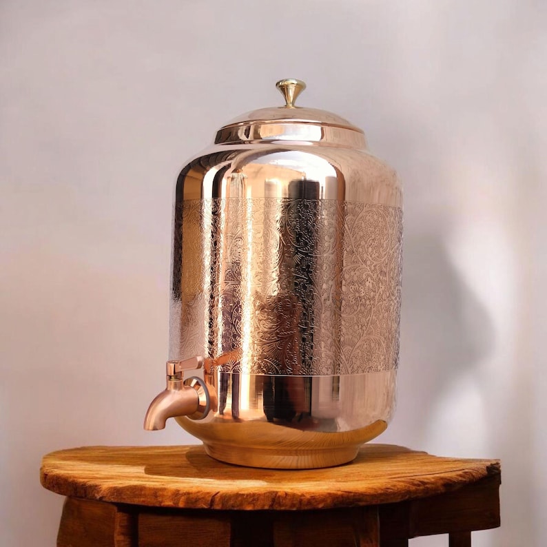 May include: A copper-colored water dispenser with a lid and a spigot. The dispenser has an engraved floral pattern on the body. The dispenser is sitting on a wooden table.