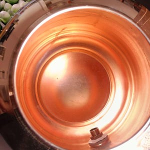 May include: A copper-colored metal container with a threaded fitting at the base. The interior of the container reflects the light, showing concentric circles. The container's smooth, polished surface gives it a clean, modern look. The container is likely used for storage or as a decorative item.