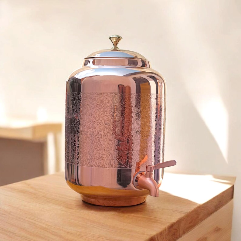 May include: A copper-colored beverage dispenser with a lid and spigot. The dispenser has an ornate floral pattern etched into the metal. It sits on a wooden base and has a small, decorative handle on top.