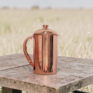 May include: A copper-coloured mug with a lid and handle. The cylindrical mug is placed on a weathered wooden surface. The background shows a blurred view of a grassy field.