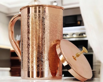 May include: A cylindrical copper pitcher with a polished finish and an intricate, embossed floral pattern covering the exterior. It features a curved handle on the side and a matching circular copper lid with a small, gold-toned metal knob handle.