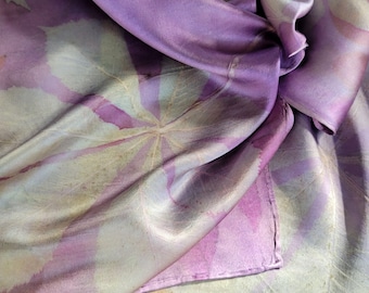 May include: A soft, purple silk scarf with a delicate floral pattern in shades of beige and pink. The scarf is folded and draped on a textured surface.