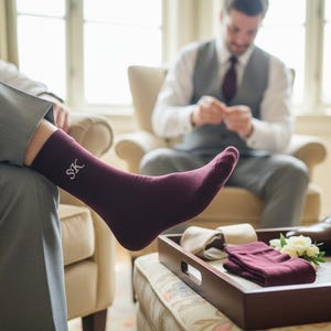 May include: A person's leg is shown wearing a burgundy sock embroidered with the initials "SK". The sock is paired with grey trousers. In the background, a man in a grey waistcoat and white shirt is seated. A tray holds socks, flowers, and brown leather shoes.