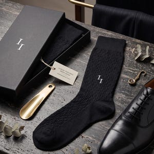 Puede incluir: Calcetines de vestir negros texturizados con las iniciales "II" bordadas en blanco, presentados en una caja negra. También se ven un calzador dorado y zapatos de vestir de cuero negro, lo que sugiere accesorios para hombre.