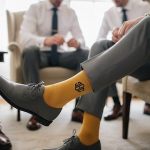 Puede incluir: Primer plano de una pierna con pantalones grises y calcetines amarillo mostaza con un monograma negro. La persona lleva zapatos de vestir grises. Hay otras personas en el fondo.