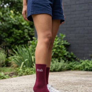 Op de afbeelding: Bordeauxkleurige sokken met de naam "Noah" in het wit geborduurd. Ze worden gedragen met een marineblauwe korte broek en witte sneakers. De persoon staat op een betonnen ondergrond met groen op de achtergrond.