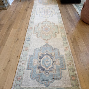 Op de afbeelding: Een lange, rechthoekige vloerkleed met een ingewikkeld geometrisch patroon in tinten lichtblauw, beige en groen. Het vloerkleed heeft een centraal ontwerp met herhalende motieven en een rand. De tekst "UrbanRugsWorld" is zichtbaar op het vloerkleed.