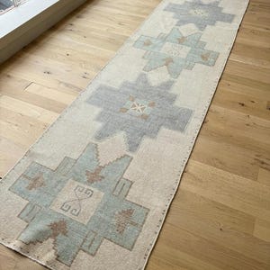 May include: A long, rectangular rug with a cream background and geometric patterns in grey, blue, and brown. The rug is placed on a light-coloured wooden floor, and the design features repeating shapes.