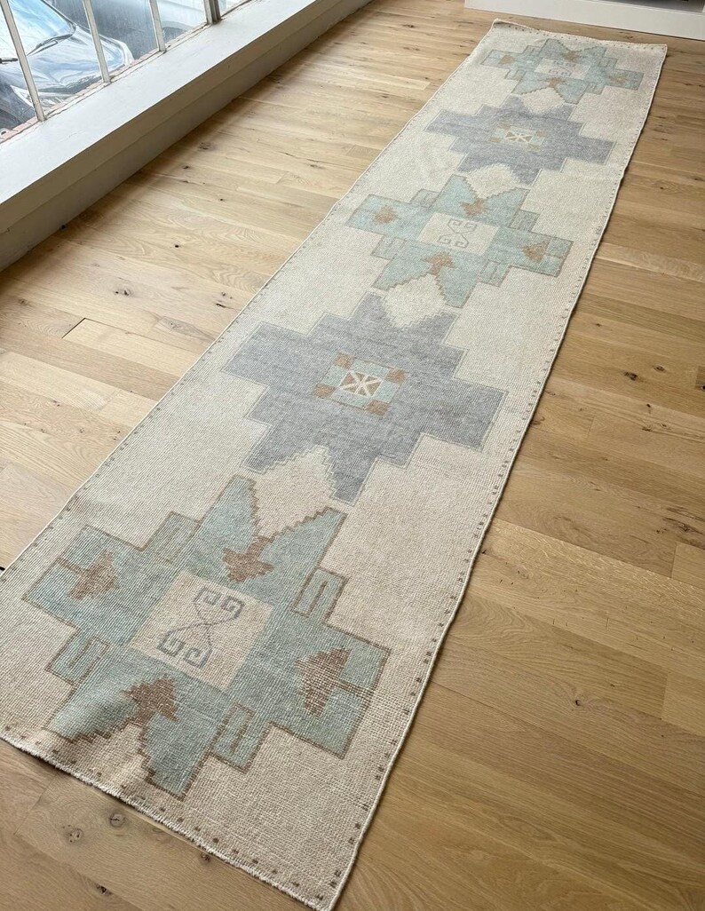May include: A long, rectangular rug with a cream background and geometric patterns in grey, blue, and brown. The rug is placed on a light-coloured wooden floor, and the design features repeating shapes.