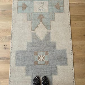 May include: A rectangular, vintage-style rug with a geometric pattern in shades of beige, grey, and light blue. The rug features a central design with symmetrical shapes and borders. The rug is placed on a wooden floor, with a person's feet wearing black studded shoes visible.