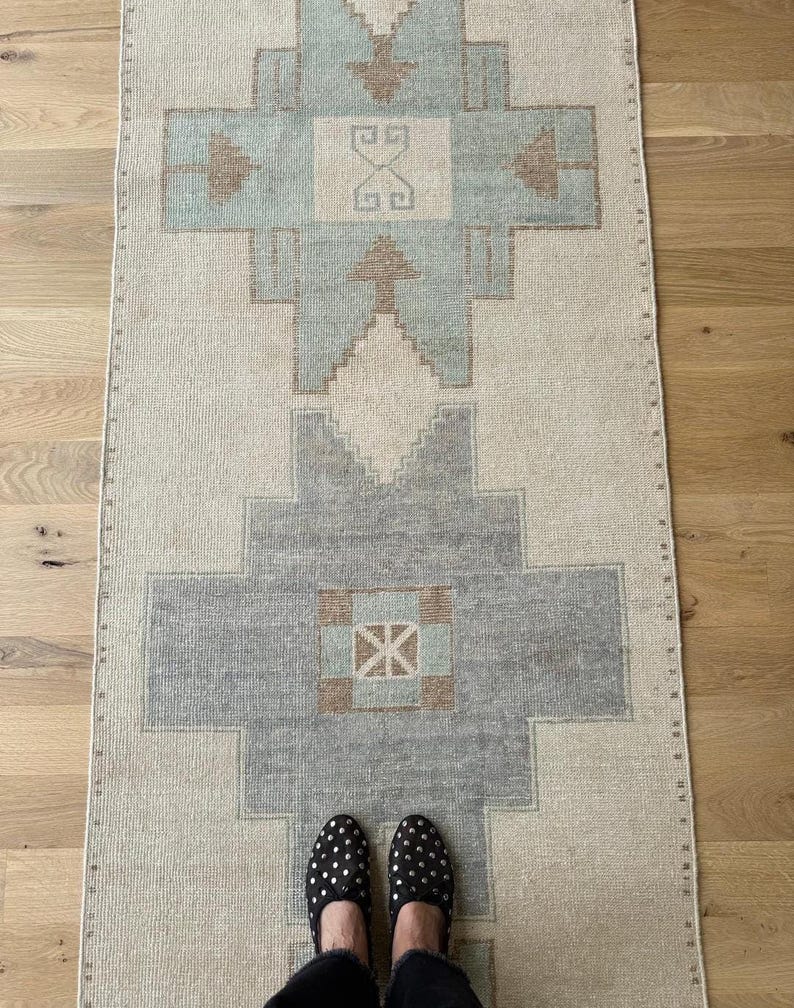 May include: A rectangular, vintage-style rug with a geometric pattern in shades of beige, grey, and light blue. The rug features a central design with symmetrical shapes and borders. The rug is placed on a wooden floor, with a person's feet wearing black studded shoes visible.